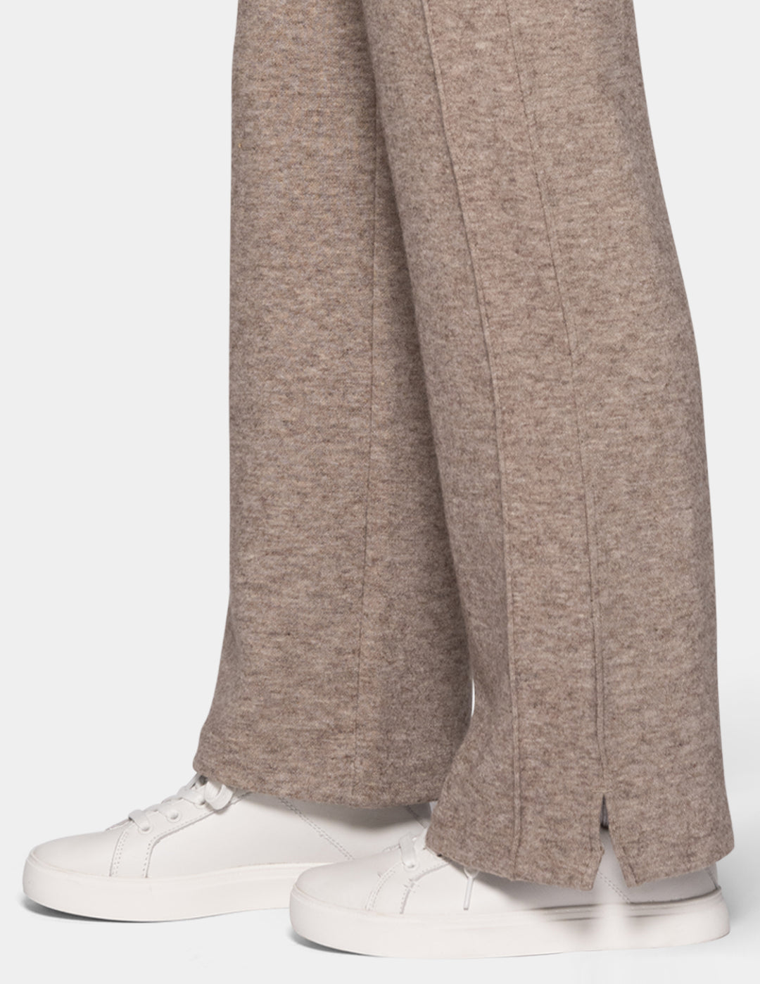 A close-up of a model wearing beige trackpants paired with white sneakers against a light grey background.