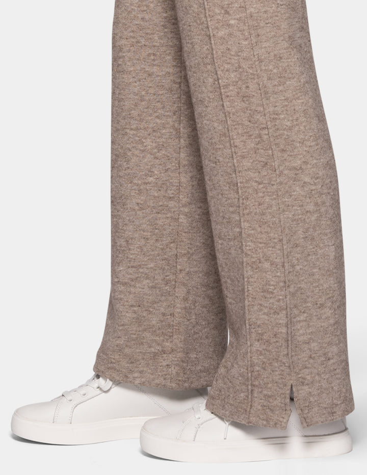 A close-up of a model wearing beige trackpants paired with white sneakers against a light grey background.