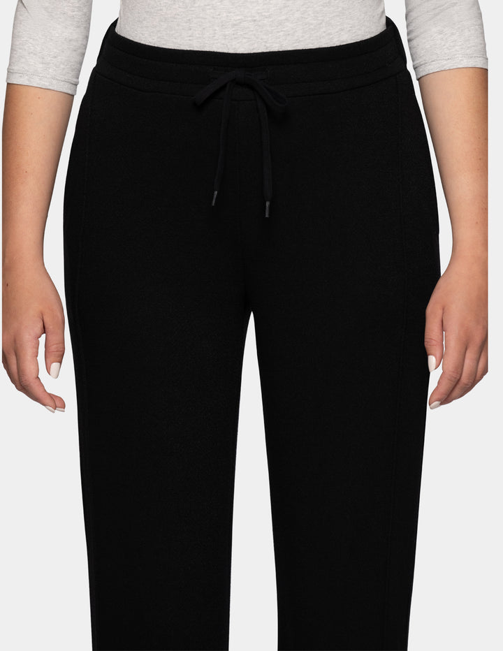 A close-up of a model wearing black jersey trackpants against a gray background.