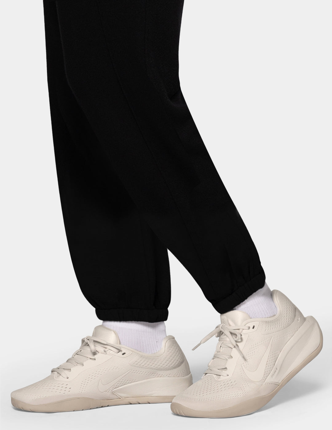 A close-up of a model wearing black jersey trackpants and off white sneakers against a gray background.