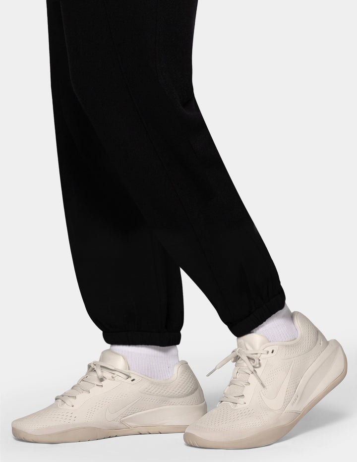 A close-up of a model wearing black jersey trackpants and off white sneakers against a gray background.