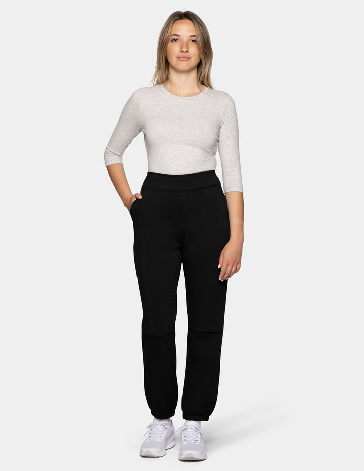 A model wearing black hi-rise joggers with a light grey scoop neck top and white sneakers against a gray background.
