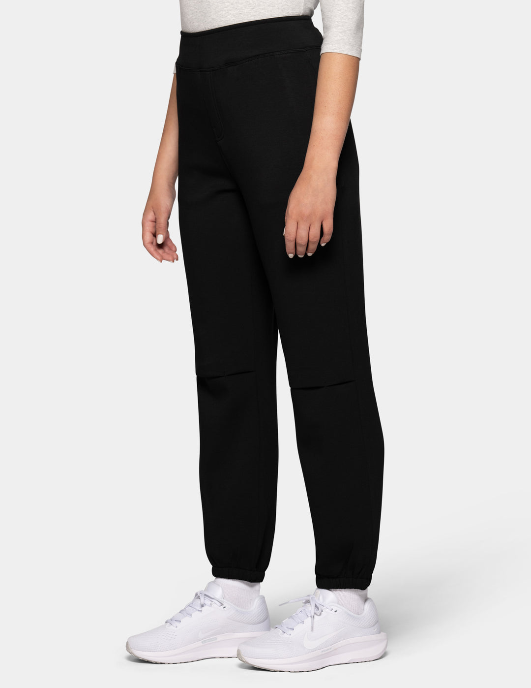 A sideview of a model wearing black hi-rise joggers and white sneakers against a gray background.
