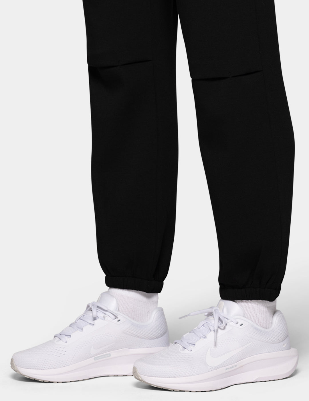 A close-up of a model wearing black hi-rise joggers and white sneakers against a gray background.