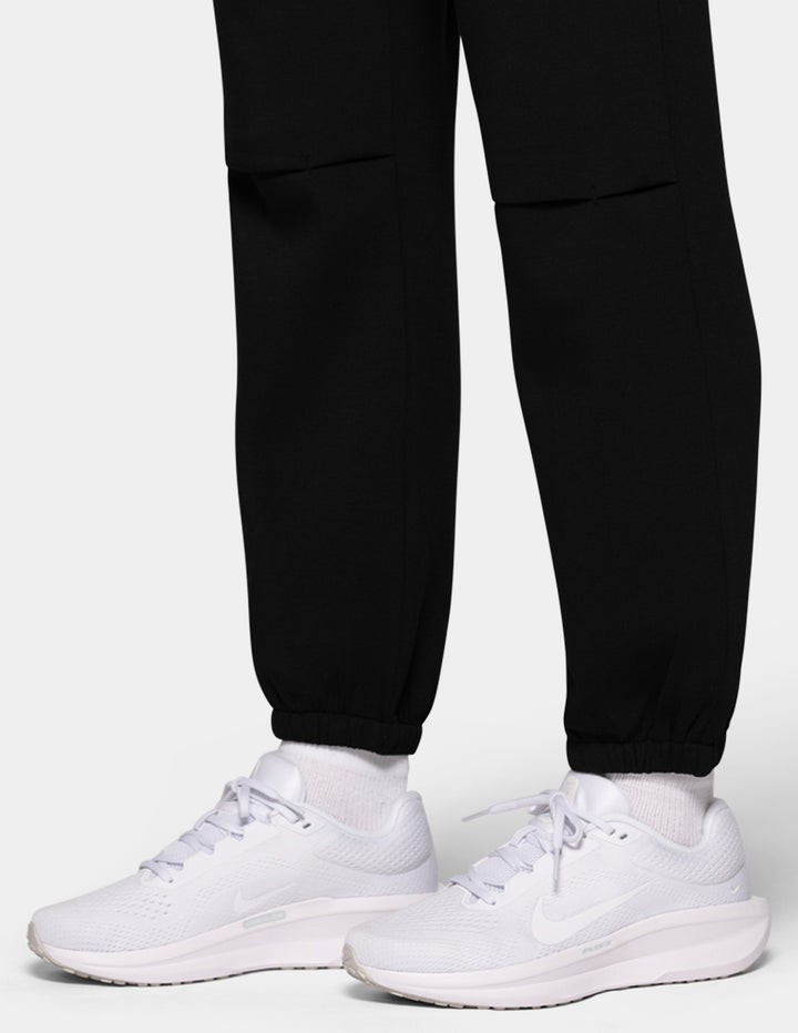 A close-up of a model wearing black hi-rise joggers and white sneakers against a gray background.