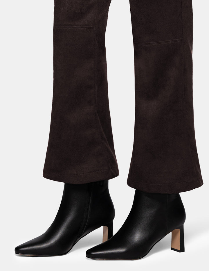 A model wearing dark brown suede flare pants paired with black boots, photographed against a light grey background.
