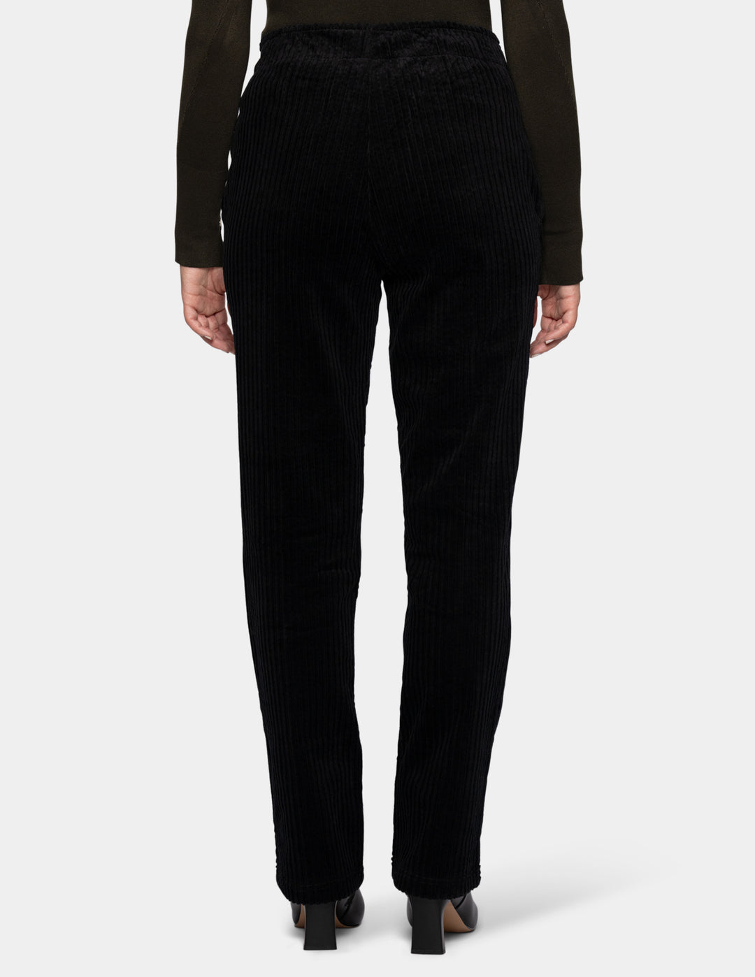 A backside view of a model wearing black corduroy pants, an olive green long sleeve top, and black boots.