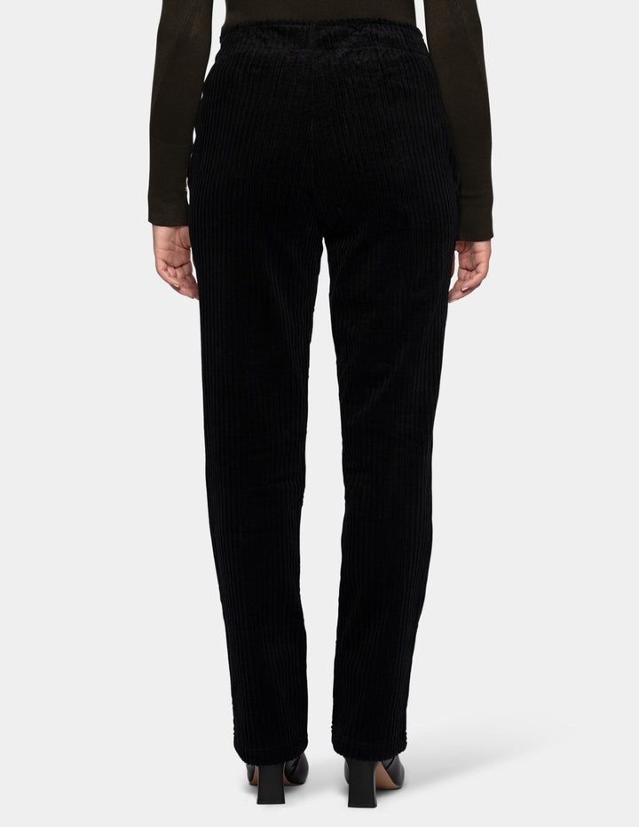 A backside view of a model wearing black corduroy pants, an olive green long sleeve top, and black boots.