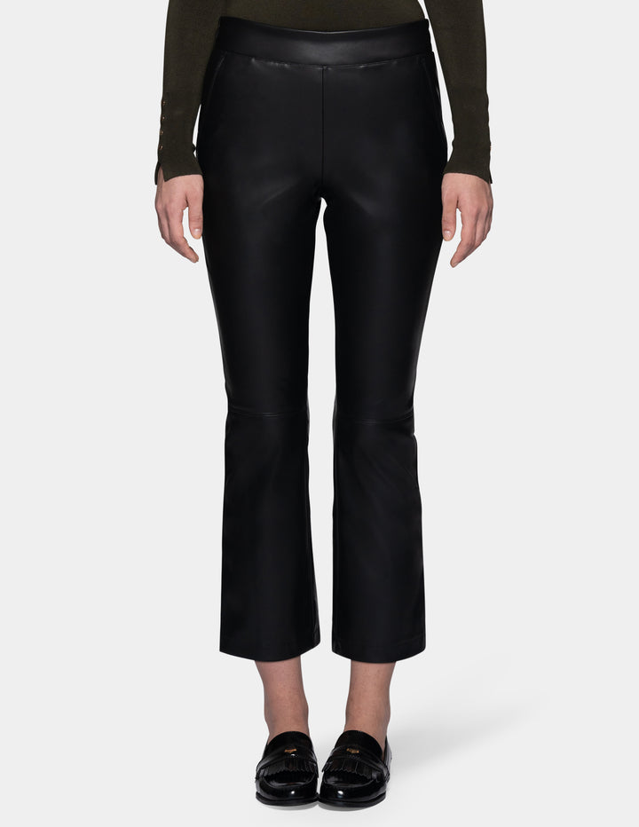 A model wearing black faux-leather cropped pants with a subtle sheen, styled with a dark olive green long-sleeve top and black loafers against a light gray studio background.