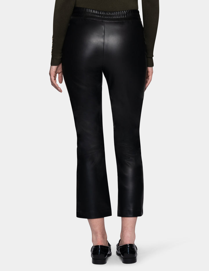 A backside view of a model wearing black faux-leather cropped pants with a subtle sheen, styled with a dark olive green long-sleeve top and black loafers against a light gray studio background.