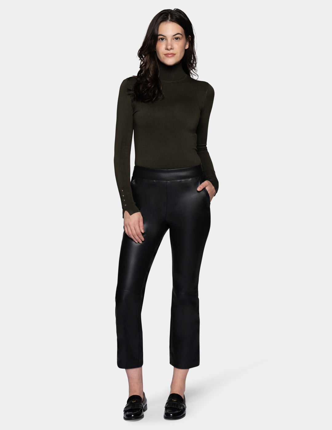 A model wearing black faux-leather cropped pants with a subtle sheen, styled with a dark olive green long-sleeve top and black loafers against a light gray studio background.
