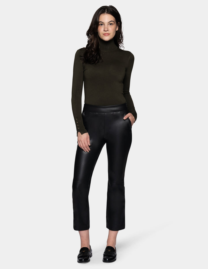 A model wearing black faux-leather cropped pants with a subtle sheen, styled with a dark olive green long-sleeve top and black loafers against a light gray studio background.