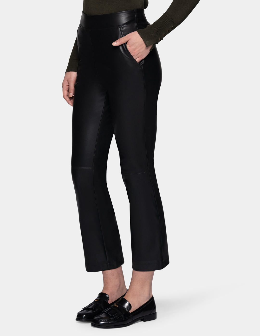 A sideview of a model wearing black faux-leather cropped pants with a subtle sheen, styled with a dark olive green long-sleeve top and black loafers against a light gray studio background.