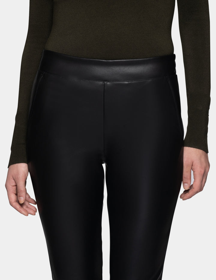 A close-up of a model wearing black faux-leather cropped pants with a subtle sheen, styled with a dark olive green long-sleeve top against a light gray studio background.