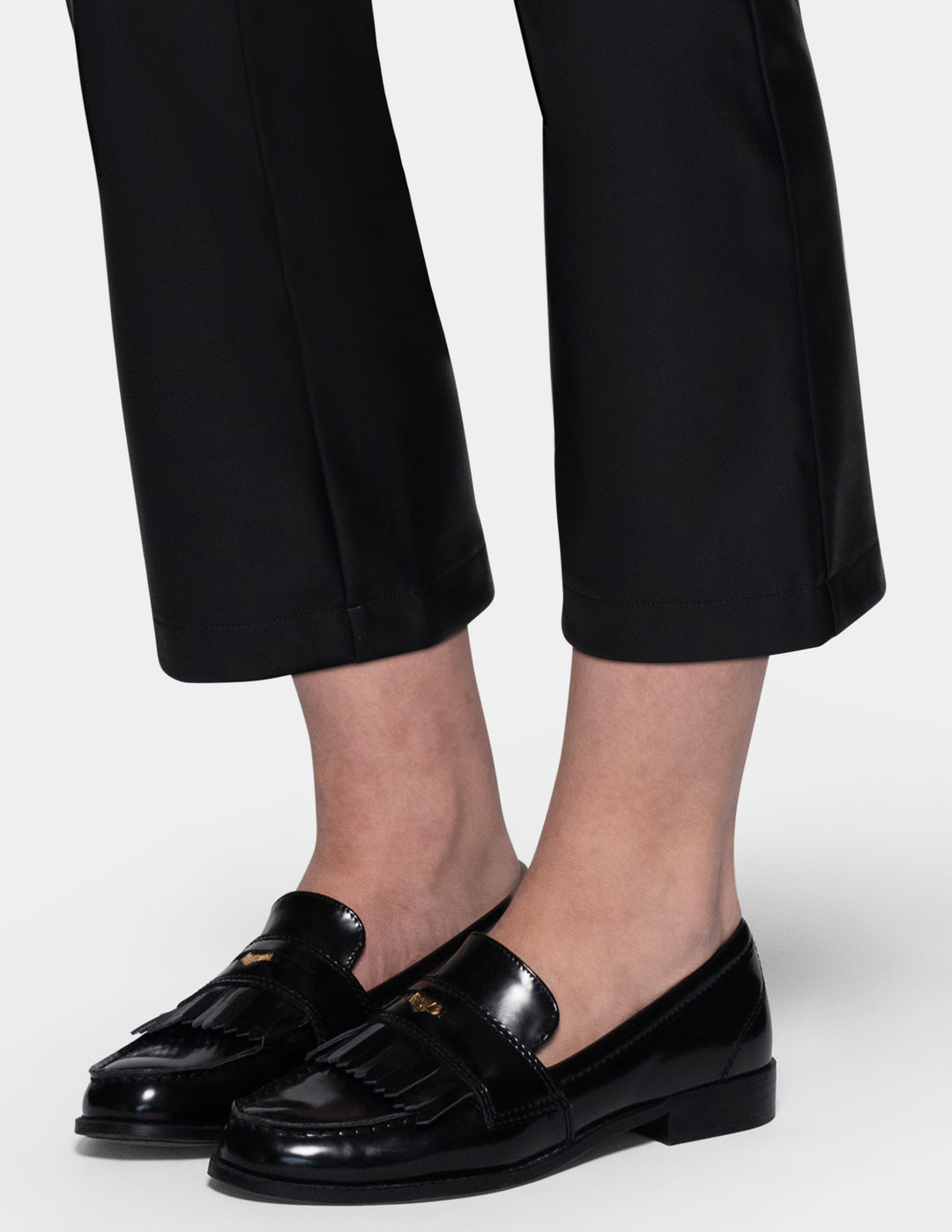 A close-up of a model wearing black faux-leather cropped pants with a subtle sheen, styled with black loafers against a light gray studio background.