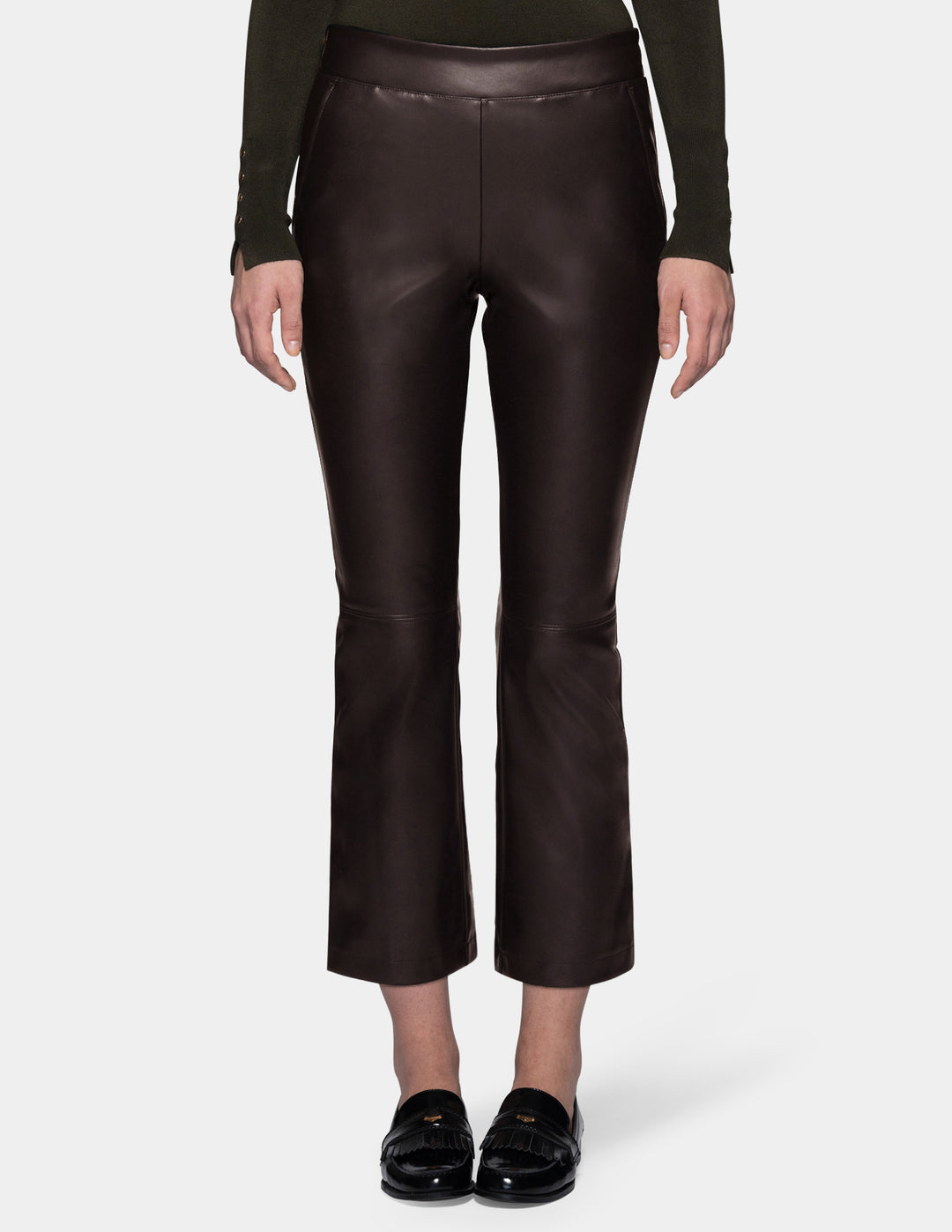 A model wearing dark brown faux-leather cropped pants with a subtle sheen, styled with a dark olive green long-sleeve top and black loafers against a light gray studio background.