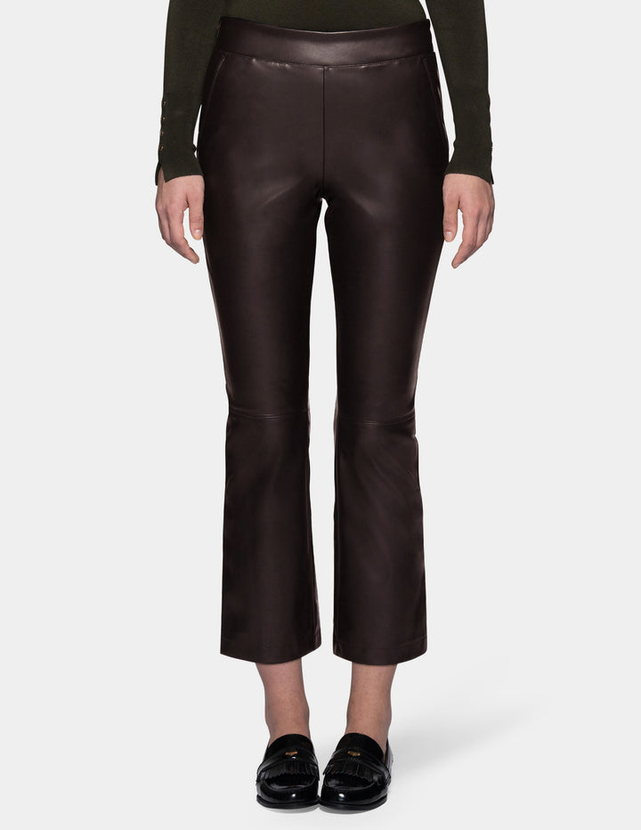 A model wearing dark brown faux-leather cropped pants with a subtle sheen, styled with a dark olive green long-sleeve top and black loafers against a light gray studio background.