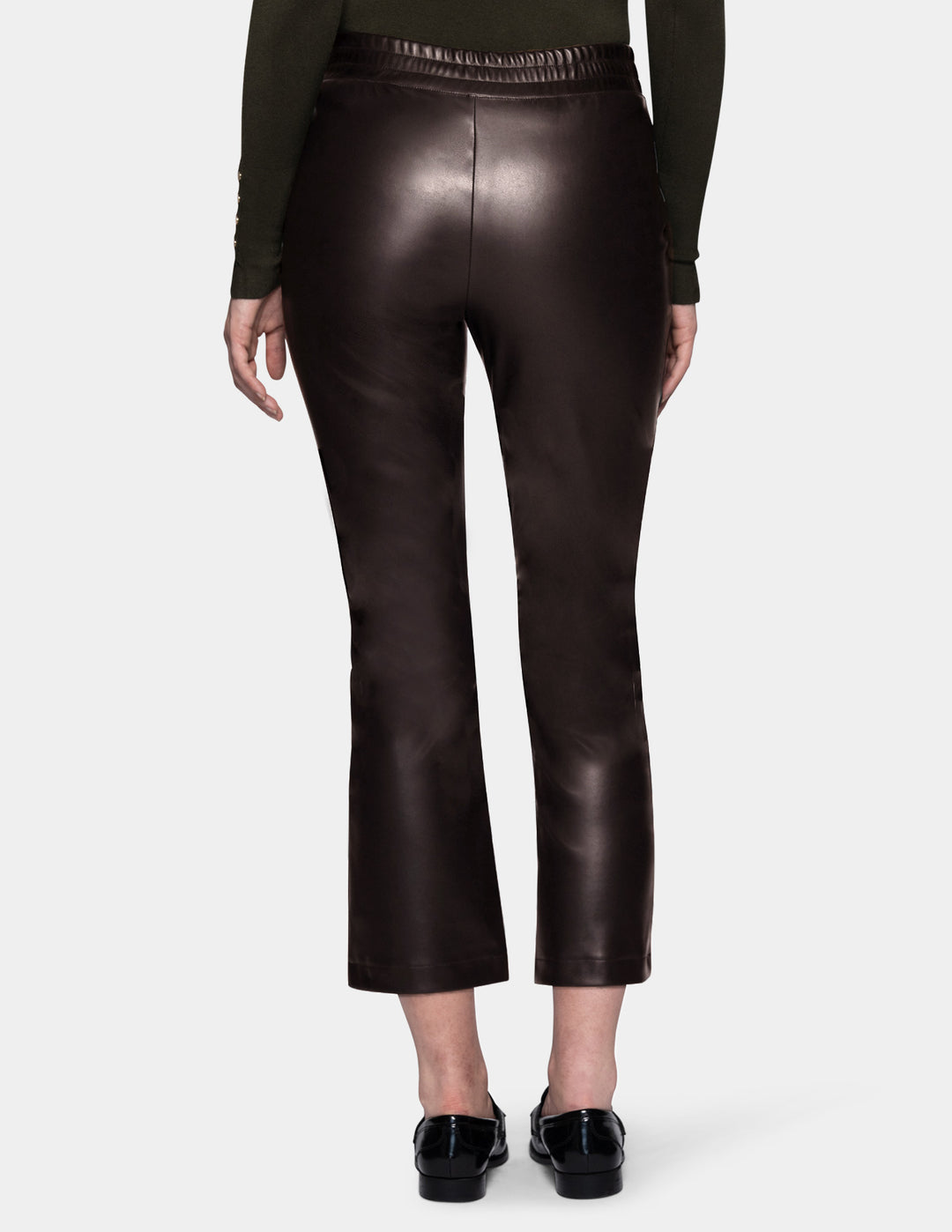 A backside view of a model wearing dark brown faux-leather cropped pants with a subtle sheen, styled with a dark olive green long-sleeve top and black loafers against a light gray studio background.
