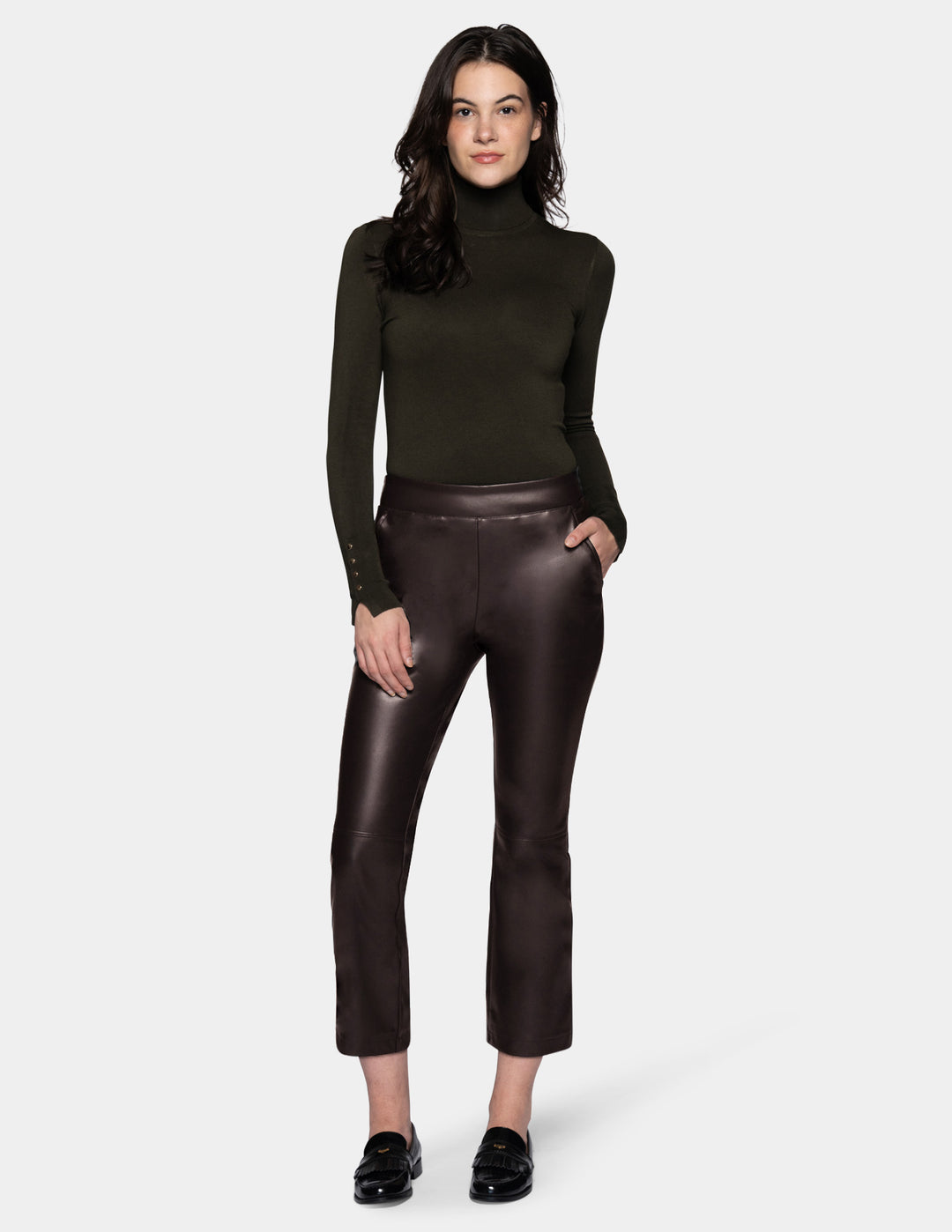 A model wearing dark brown faux-leather cropped pants with a subtle sheen, styled with a dark olive green long-sleeve top and black loafers against a light gray studio background.