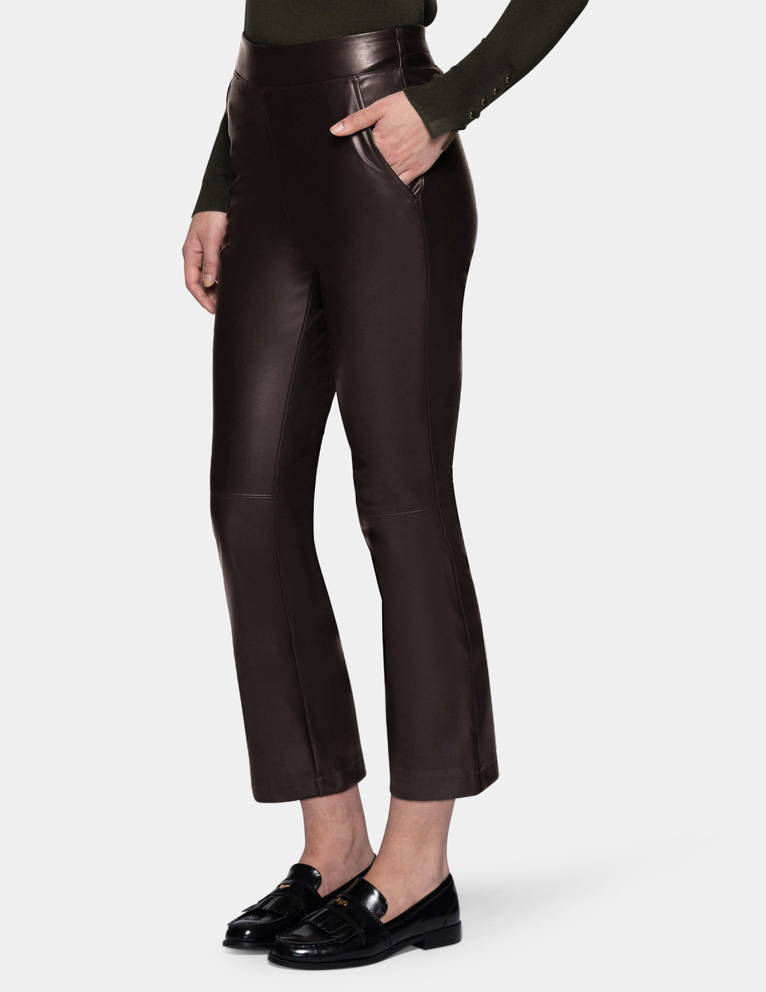 A model wearing dark brown faux-leather cropped pants with a subtle sheen, styled with a dark olive green long-sleeve top and black loafers against a light gray studio background.
