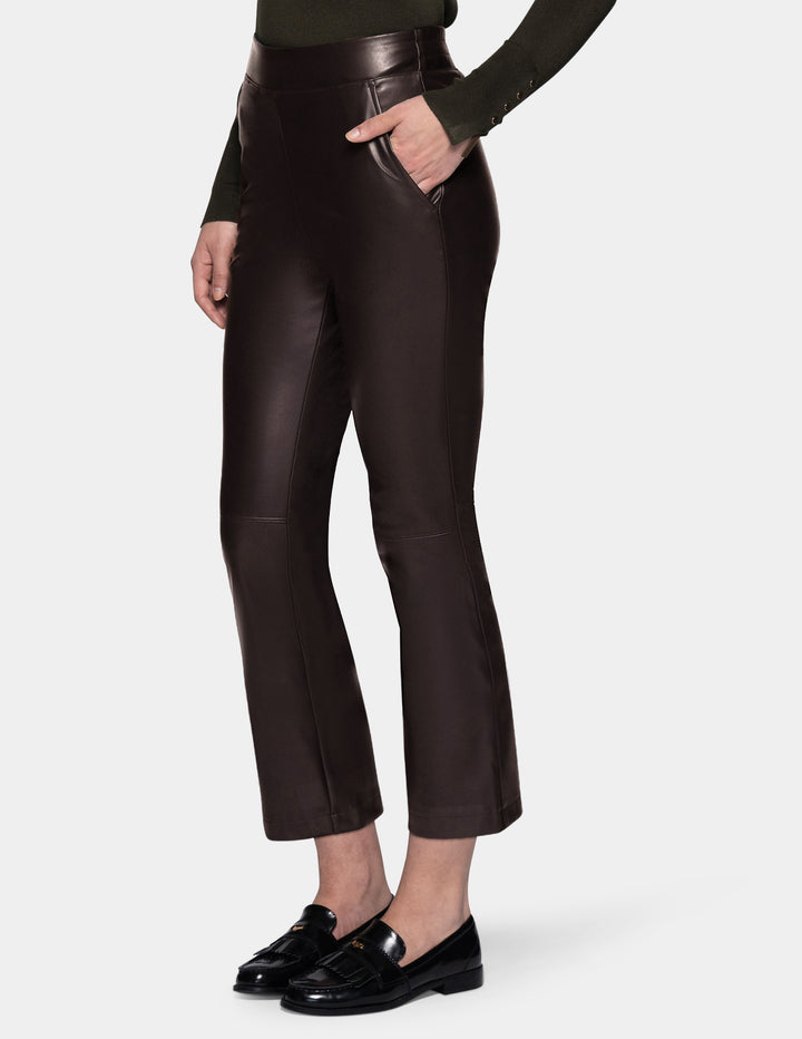A model wearing dark brown faux-leather cropped pants with a subtle sheen, styled with a dark olive green long-sleeve top and black loafers against a light gray studio background.