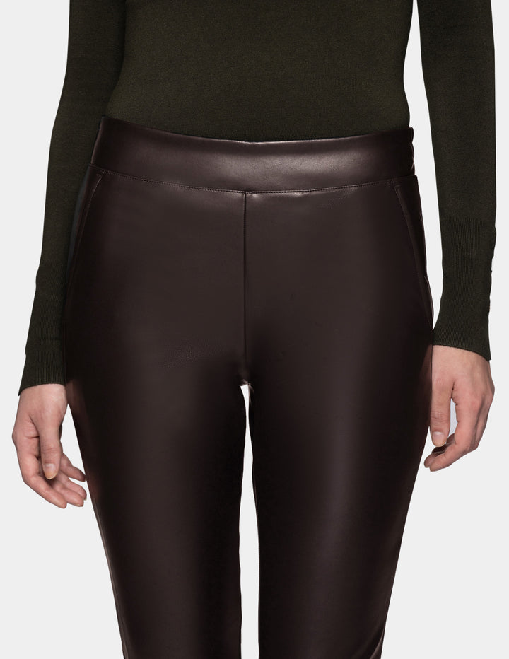 A person wearing dark brown faux-leather leggings and an olive green long-sleeve top, shown from the chest down against a light gray studio background.