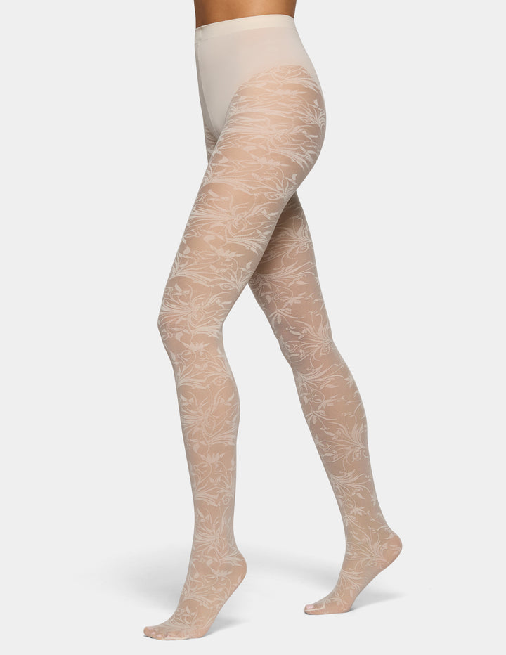 A model's legs wearing sheer nude tights with a delicate white floral pattern, shown against a light gray studio background.