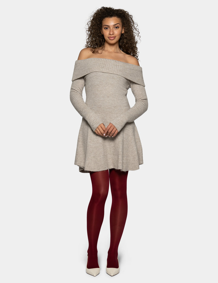 A woman modeling a light gray off-the-shoulder knit mini dress with long sleeves, paired with dark red tights and white pointed-toe heels, against a light gray background.