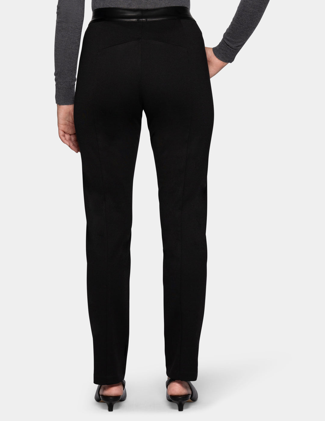 A backside view of a model wearing black faux leather pants, a long sleeve grey top, and black kitten heels.