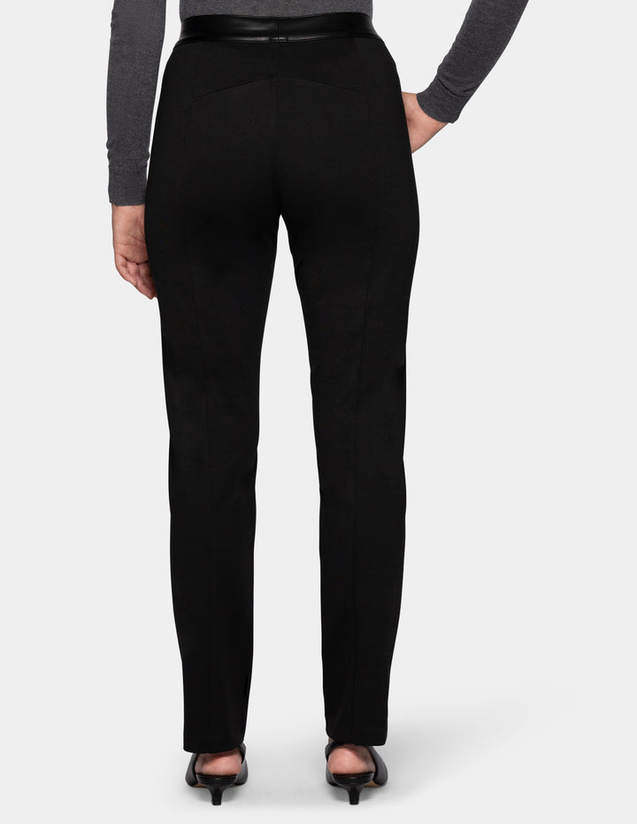A backside view of a model wearing black faux leather pants, a long sleeve grey top, and black kitten heels.