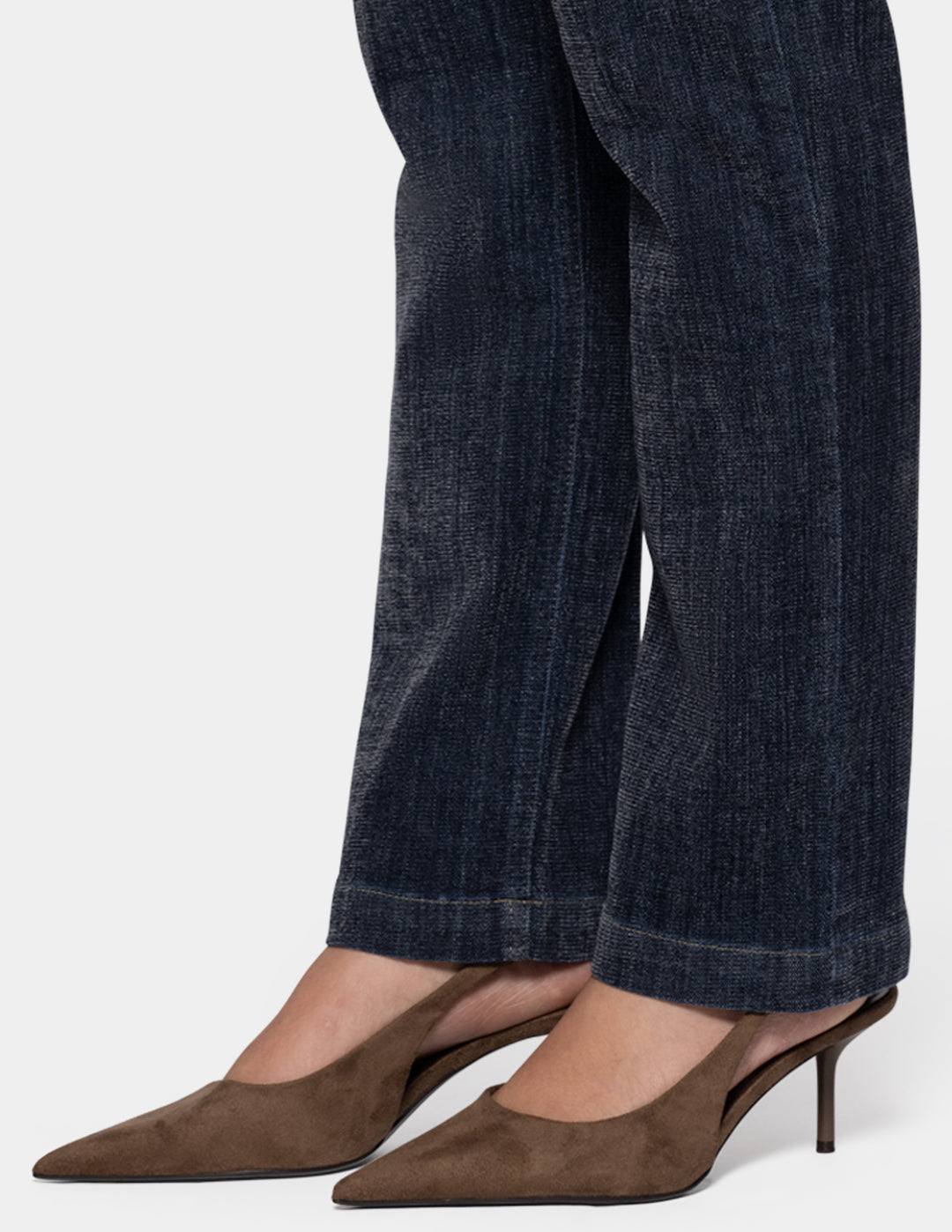 A model wearing dark navy blue denim and brown pointed-toe heels against a light gray background.