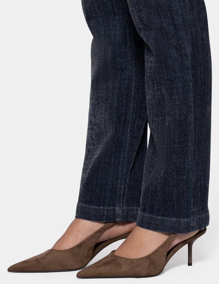 A model wearing dark navy blue denim and brown pointed-toe heels against a light gray background.