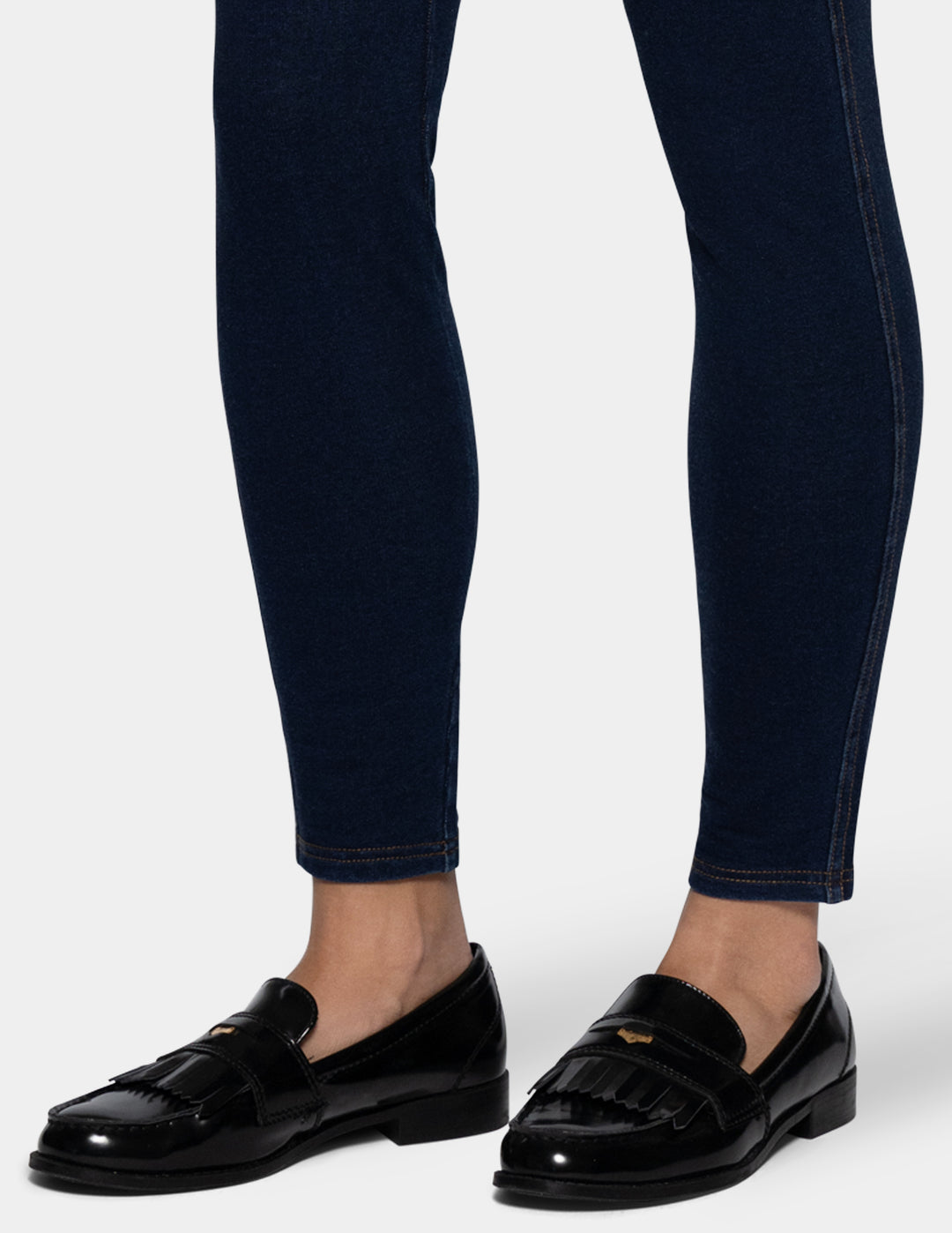 A close-up of a person's legs wearing dark navy blue leggings and shiny black patent leather tassel loafers with a small gold logo on the strap.