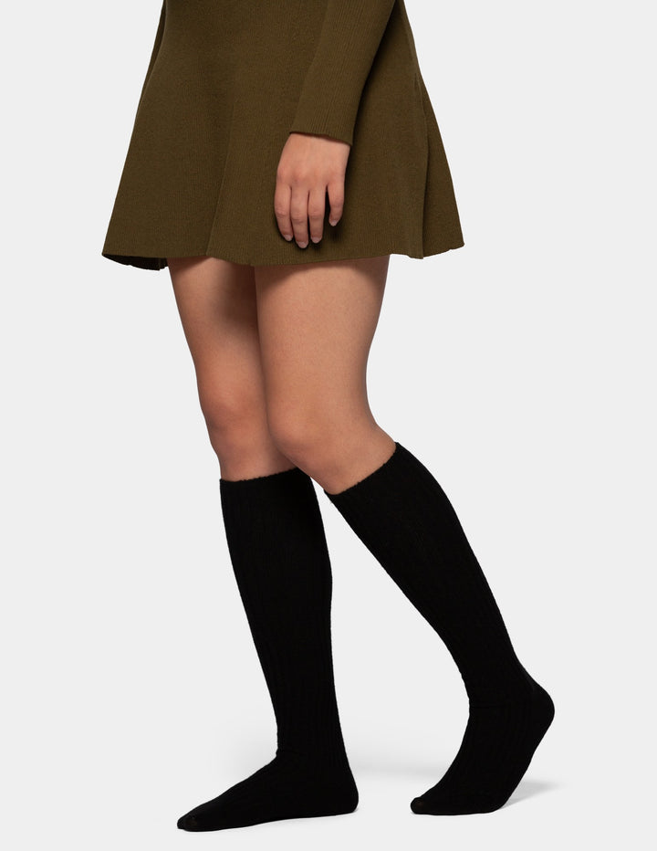 A model from the waist down wearing an olive green pleated mini dress, and black knee-high socks against a light gray studio background.