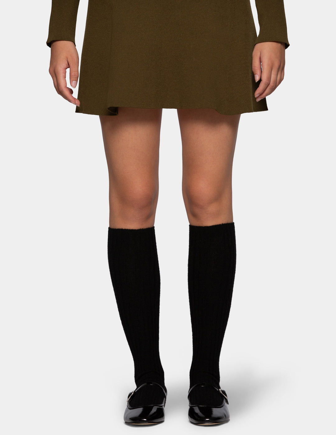 A model from the waist down wearing an olive green mini dress, black ribbed knee-high socks, and black patent leather loafers against a light gray background.