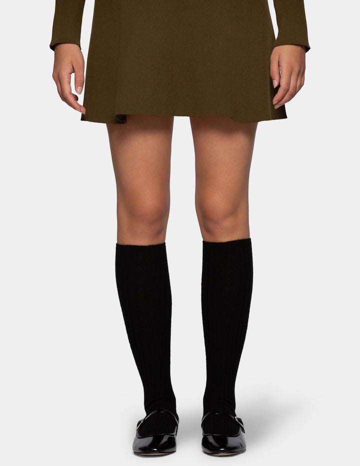 A model from the waist down wearing an olive green mini dress, black ribbed knee-high socks, and black patent leather loafers against a light gray background.