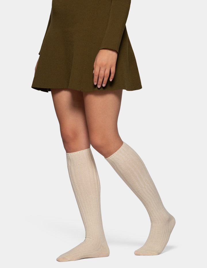A model from the waist down wearing ribbed knee-high socks in a light beige color, paired with an olive green skirt, against a light gray studio background.