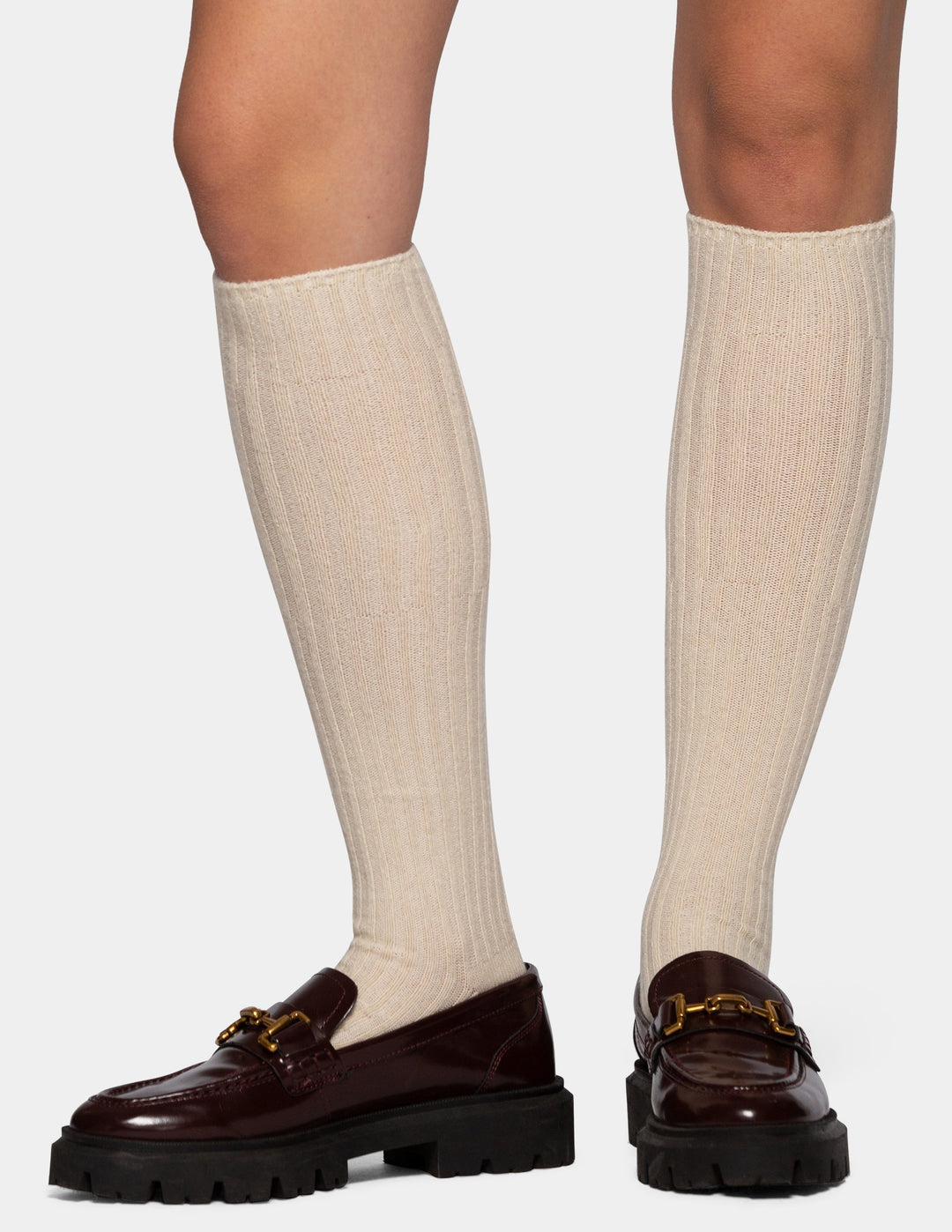 A person's legs wearing ribbed cream knee-high socks and dark brown leather loafers with chunky black soles and gold-tone hardware, shown against a light gray studio background.