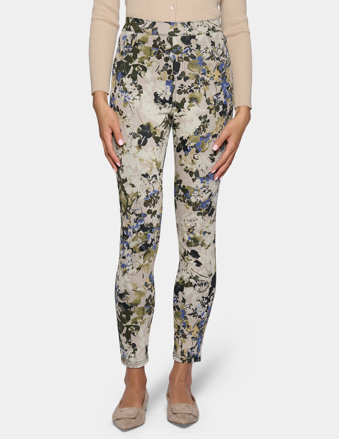 A model wearing high-waisted, slim-fit ankle-length pants with a dense floral and botanical print. Paired with a beige long-sleeve top and beige pointed-toe flats, set against a plain light gray studio background.