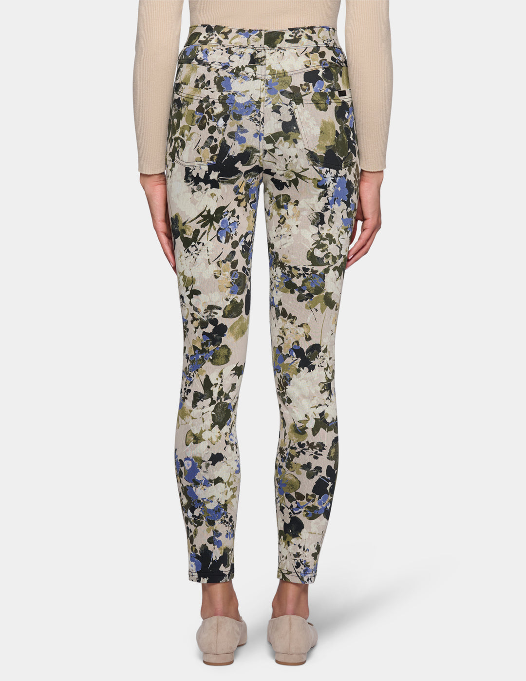 A backside view of a model wearing high-waisted, slim-fit ankle-length pants with a dense floral and botanical print. Paired with a beige long-sleeve top and beige pointed-toe flats, set against a plain light gray studio background.