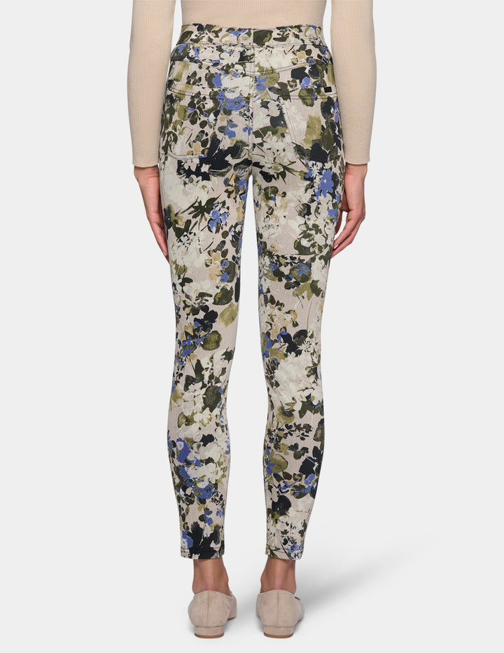 A backside view of a model wearing high-waisted, slim-fit ankle-length pants with a dense floral and botanical print. Paired with a beige long-sleeve top and beige pointed-toe flats, set against a plain light gray studio background.