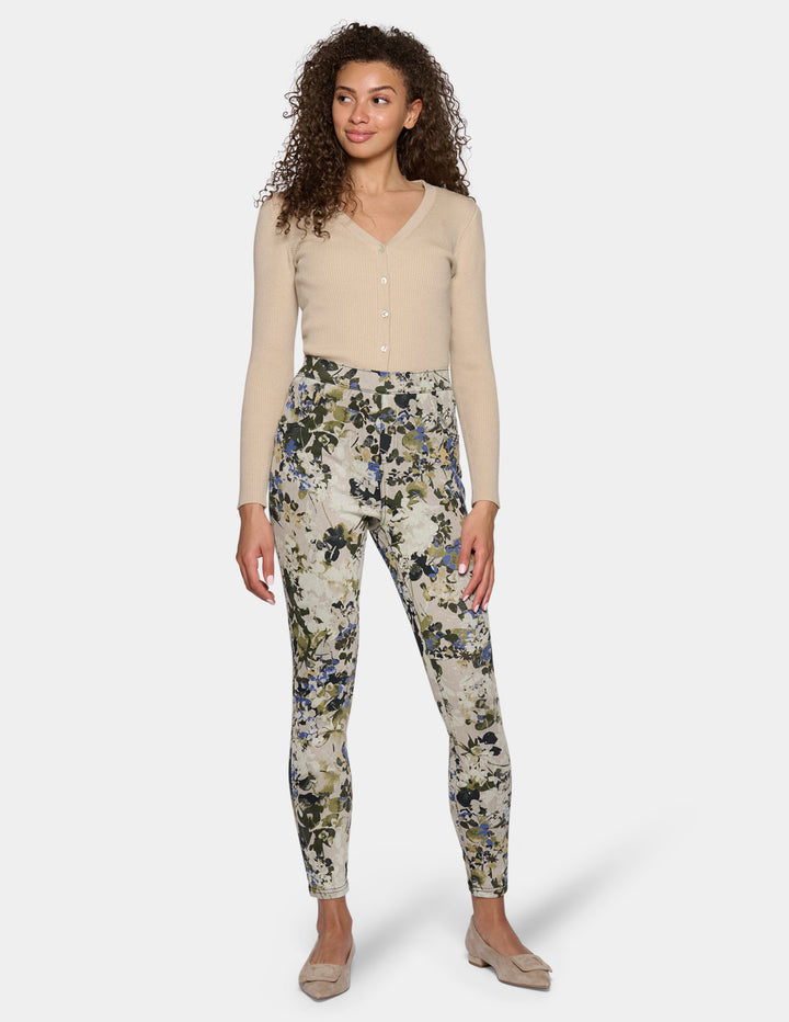 A model wearing high-waisted, slim-fit ankle-length pants with a dense floral and botanical print. Paired with a beige long-sleeve top and beige pointed-toe flats, set against a plain light gray studio background.