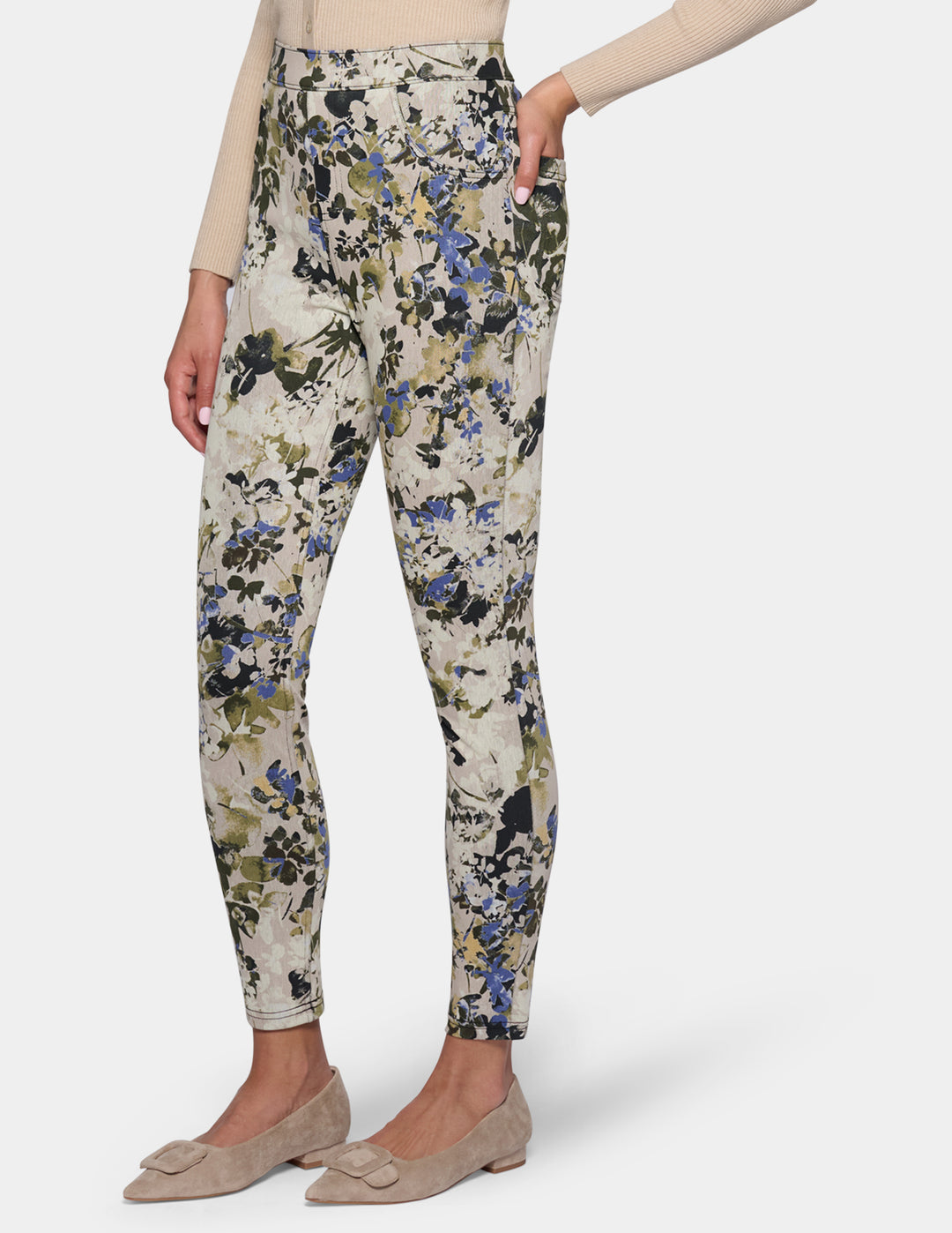 A sideview of a model wearing high-waisted, slim-fit ankle-length pants with a dense floral and botanical print. Paired with a beige long-sleeve top and beige pointed-toe flats, set against a plain light gray studio background.