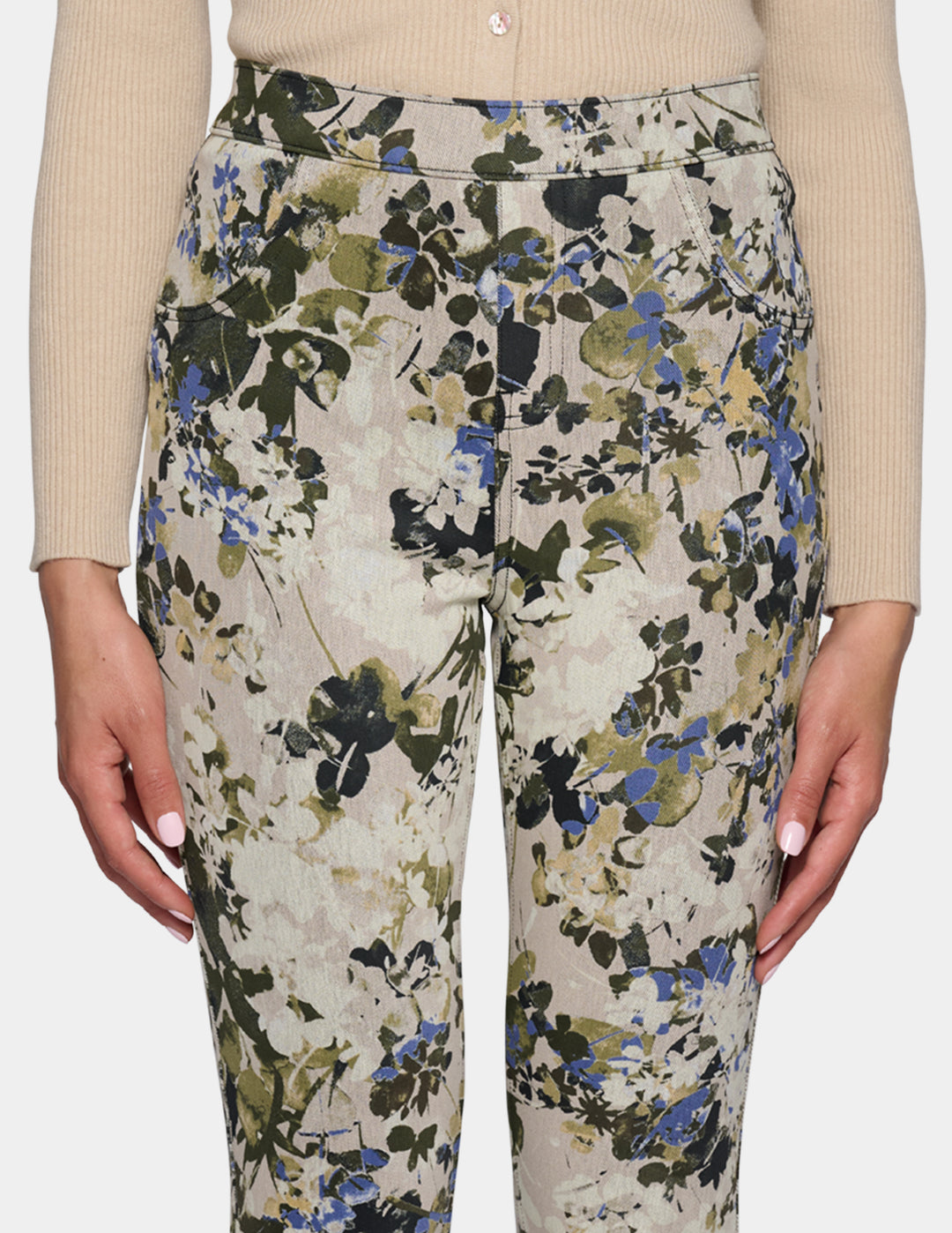 A close-up of a model wearing high-waisted, slim-fit ankle-length pants with a dense floral and botanical print. Paired with a beige long-sleeve top and beige pointed-toe flats, set against a plain light gray studio background.