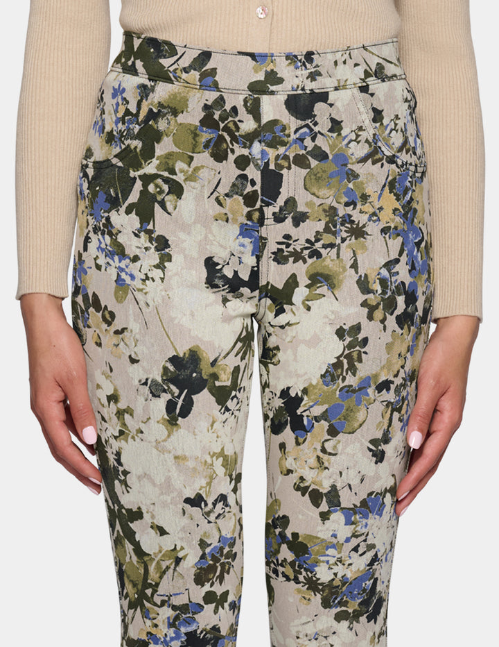 A close-up of a model wearing high-waisted, slim-fit ankle-length pants with a dense floral and botanical print. Paired with a beige long-sleeve top and beige pointed-toe flats, set against a plain light gray studio background.