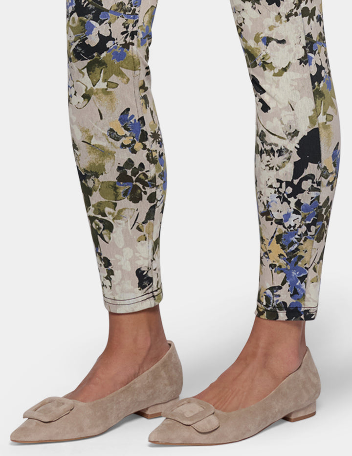 A model wearing slim-fit ankle-length pants with a dense floral and botanical print. Paired with beige pointed-toe flats, set against a plain light gray studio background.