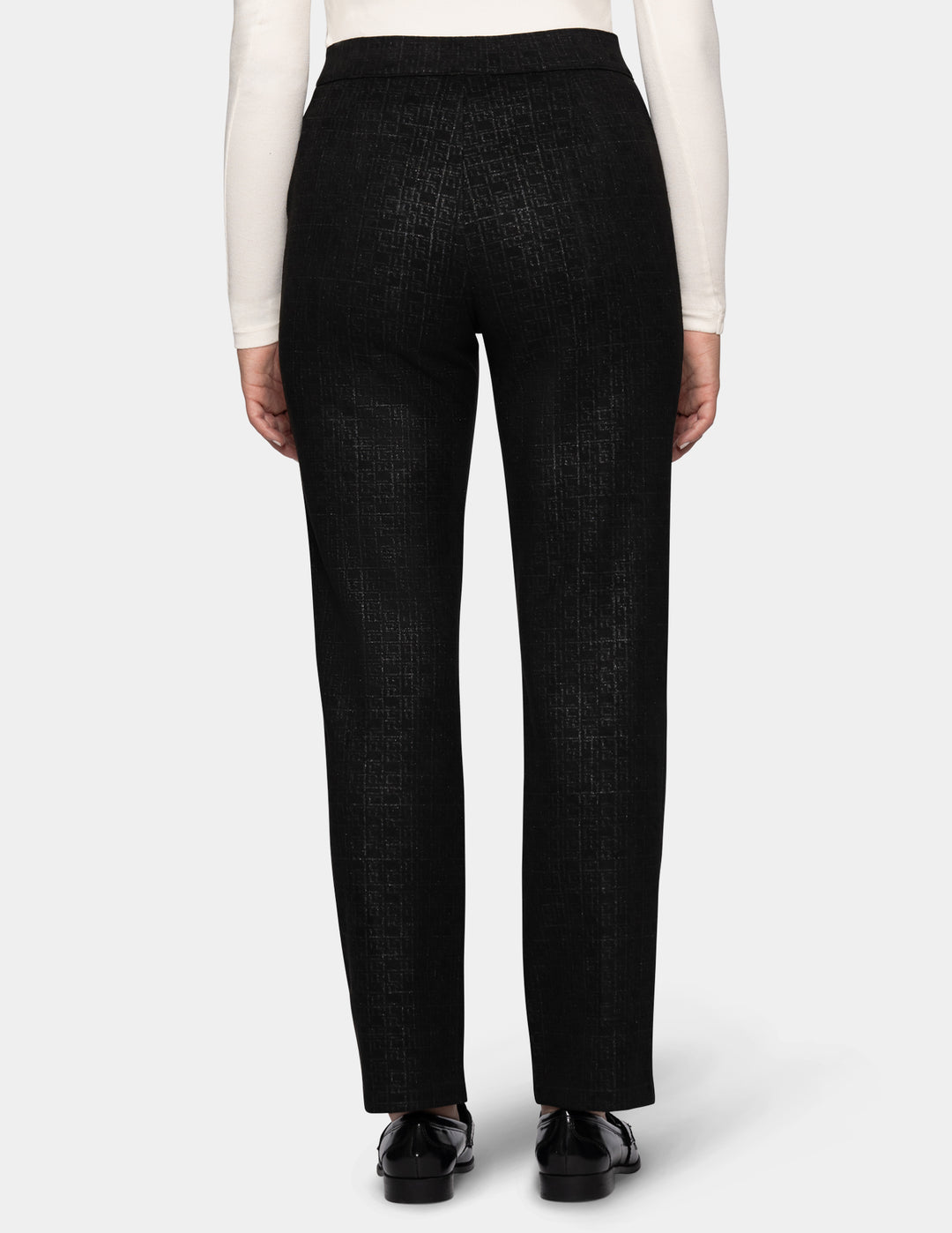 A backside view of a model wearing black textured trousers, a white long-sleeve top, and black loafers, set against a plain light gray studio background.