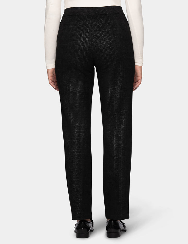 A backside view of a model wearing black textured trousers, a white long-sleeve top, and black loafers, set against a plain light gray studio background.