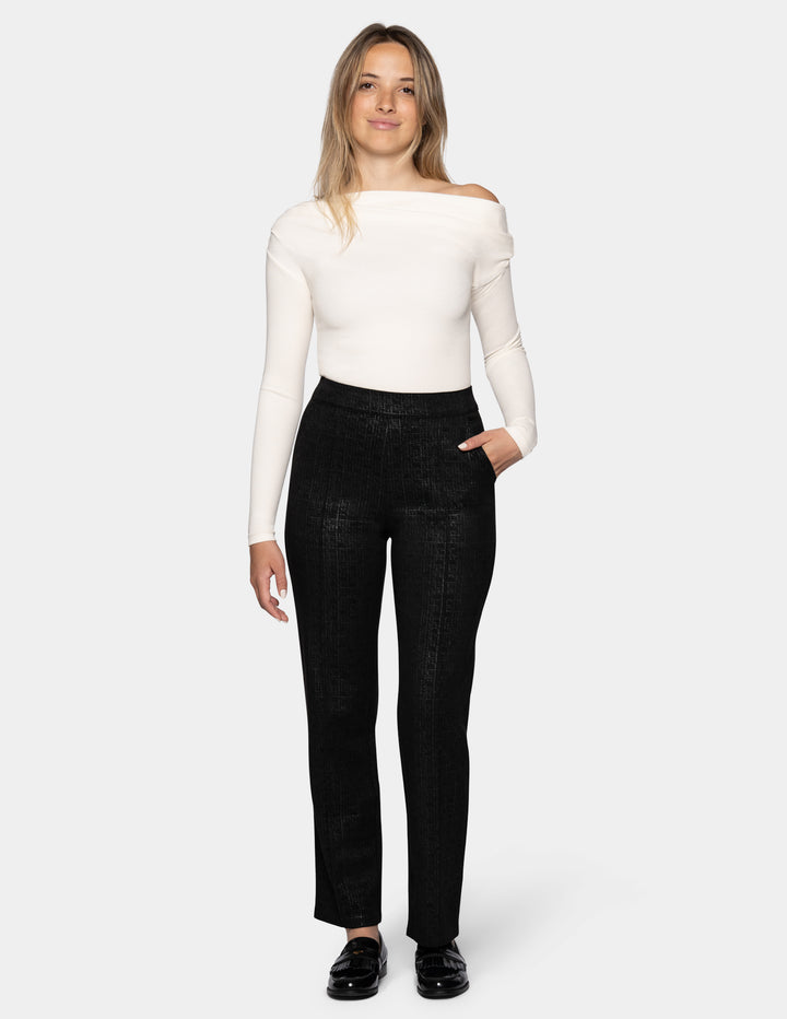 A model wearing black textured trousers, a white long-sleeve top, and black loafers, set against a plain light gray studio background.