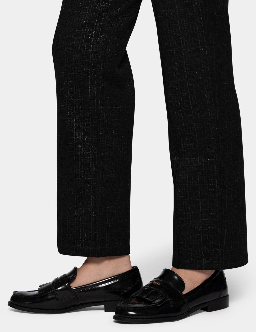 A close-up view of a model wearing black textured trousers and black loafers, set against a plain light gray studio background.